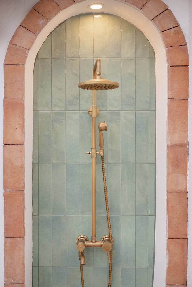 Ducha exterior de latón envejecido bajo un arco de barro y azulejo verde.