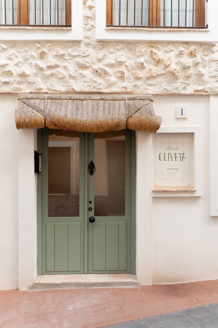 La puerta verde de dos hojas bajo la persiana de esparto, con la placa de azulejo.